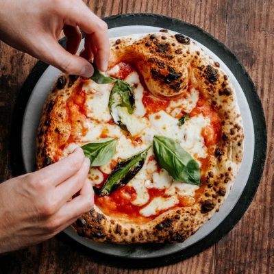 Close-up of hands adding basil to a freshly baked Neapolitan pizza, highlighting its cheesy and delicious appeal.